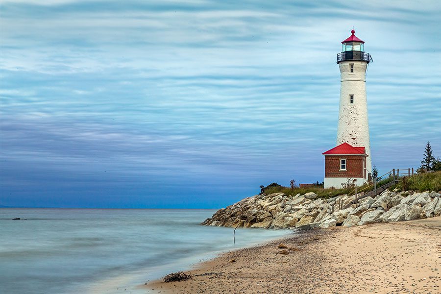 About Our Agency - Lighthouse on a Rocky Coastline by the Lake in Michigan Against a Cloudy Blue Sky at Sundown