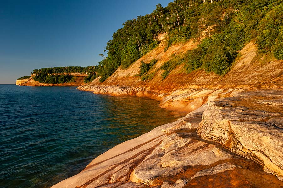Client Center - View of Rocks Along the Coastline at Lake Superior in Michigan with Green Foliage and a Clear Blue Sky at Sunset