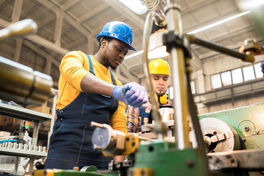 Specialized Business Insurance - Portrait of Two Young Men Wearing Hard Hats Using Machinery While Workinig Inside a Manufacturing Facility